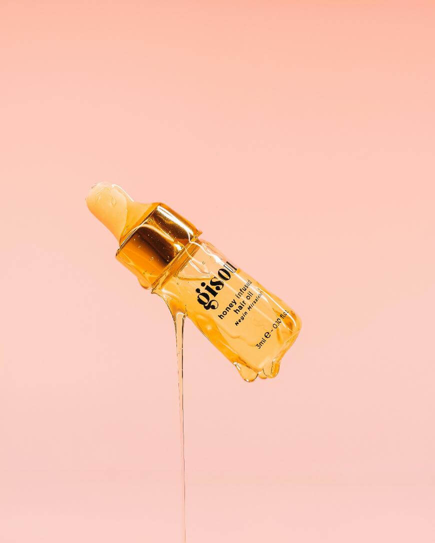 A dropper bottle of Gisou Honey Infused Hair Oil is tilted against a pink background, with golden oil dripping from the bottle.