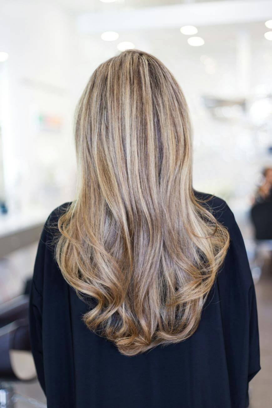 A person with long, wavy, blonde hair with highlights, wearing a black cape, is shown from behind in a brightly lit hair salon.