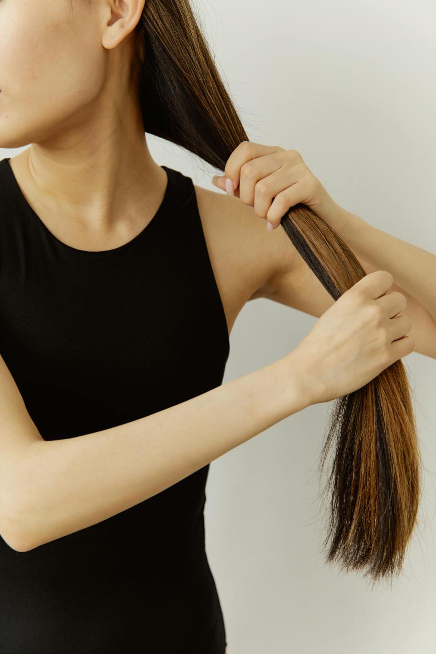A woman wearing a sleeveless black top holds and smooths her long, straight hair, which has brown highlights, against a plain light background. Her face is turned away from the camera.