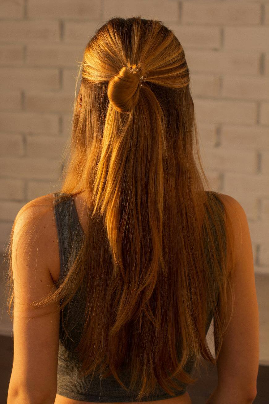 A woman with long, straight, light brown hair styled in a half-up bun stands facing away from the camera, wearing a sleeveless gray top in front of a white brick wall.