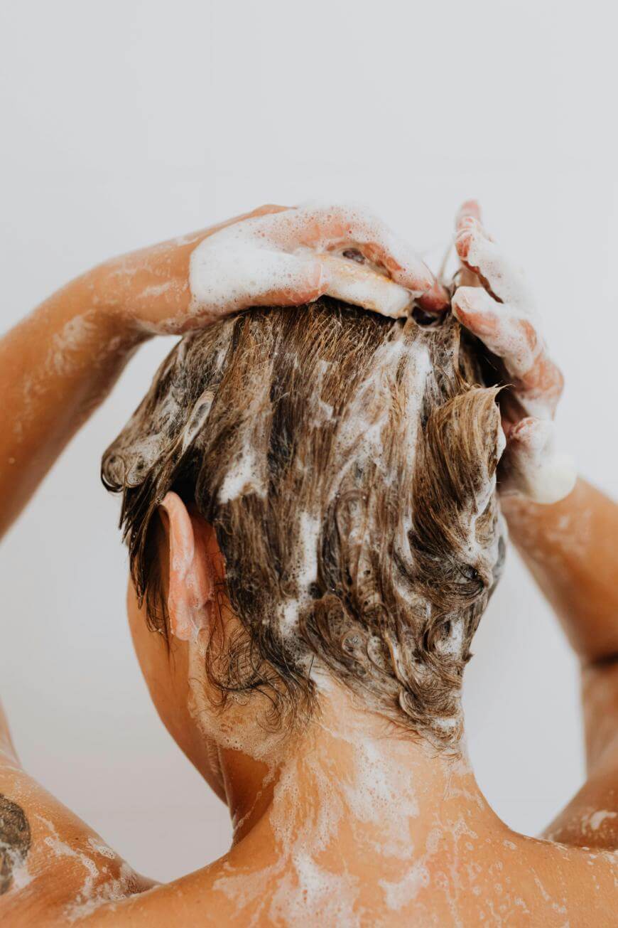 A person washing their hair, seen from behind, with their hands massaging shampoo into their short brown hair, creating a thick lather of bubbles and foam.
