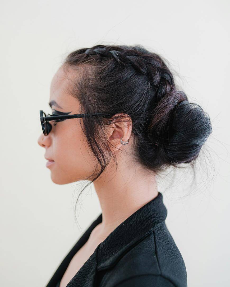 A woman in profile wears black sunglasses and a black jacket. Her dark hair is styled in a braid crown that leads into a low, messy bun. She stands against a plain, light background.