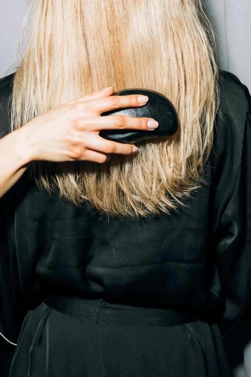 A person with long, straight blonde hair is seen from behind, wearing a black robe and brushing their hair with a black hairbrush.