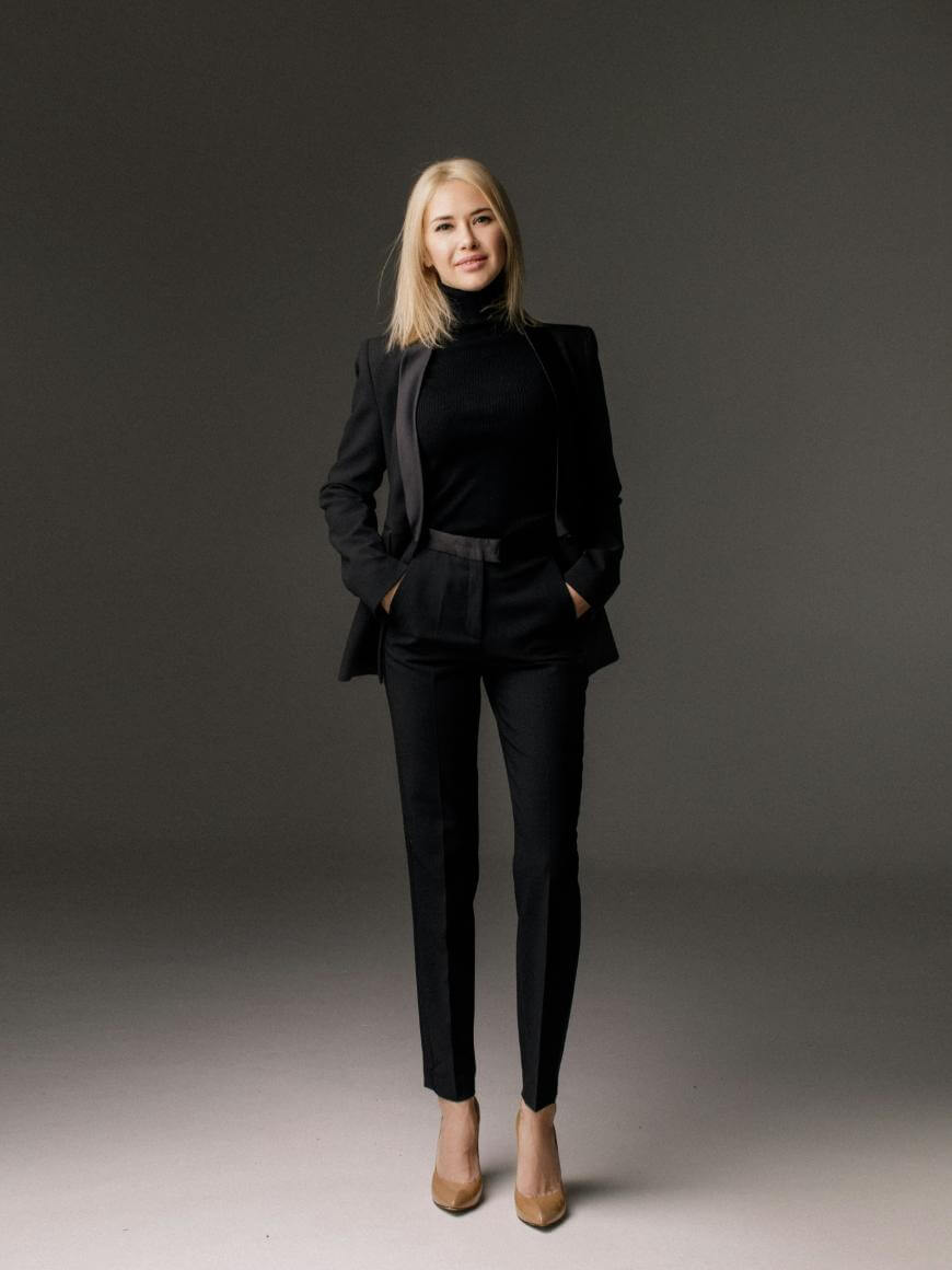 A woman with straight blonde hair stands against a plain gray background, wearing a black suit with a black turtleneck and tan high heels. She has her hands in her pockets and is smiling slightly.