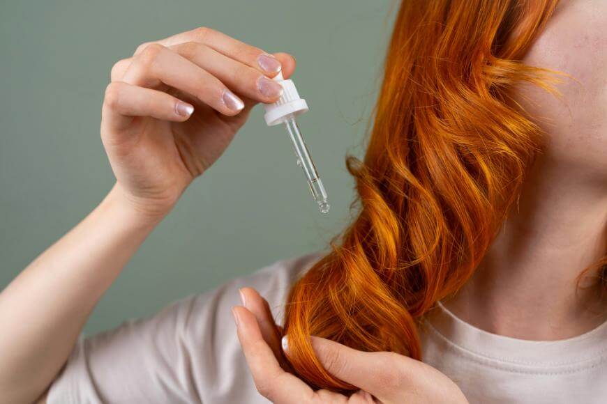 A person with long, wavy red hair applies oil or serum to their hair using a dropper, against a plain light green background. Their face is partially out of frame.