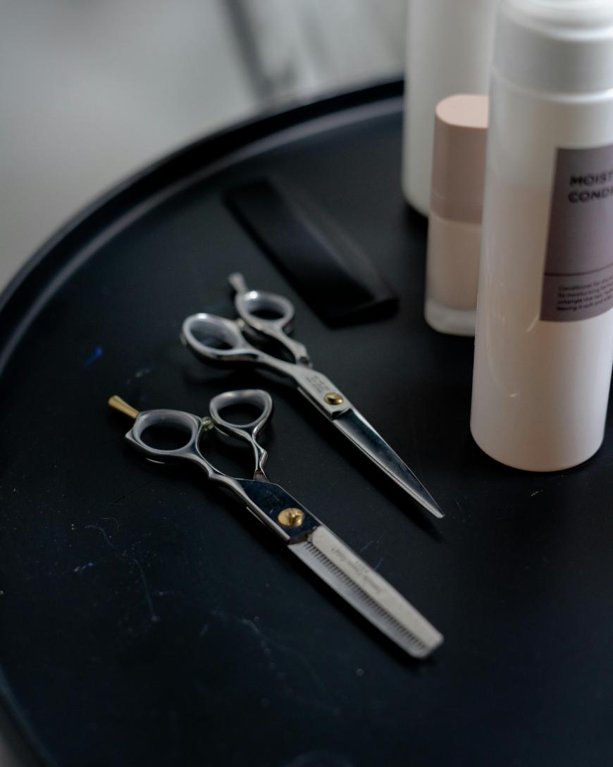 Two pairs of hairdressing scissors, a black comb, and bottles of hair products are arranged on a round black tray.