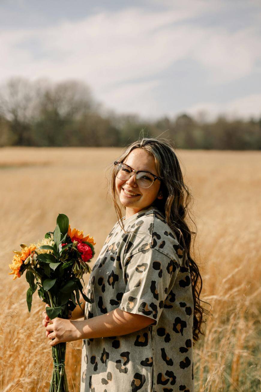 A woman wearing glasses and a leopard-print dress stands in a golden field, smiling at the camera and holding a bouquet of colorful flowers. Trees and a blue sky with clouds are visible in the background.