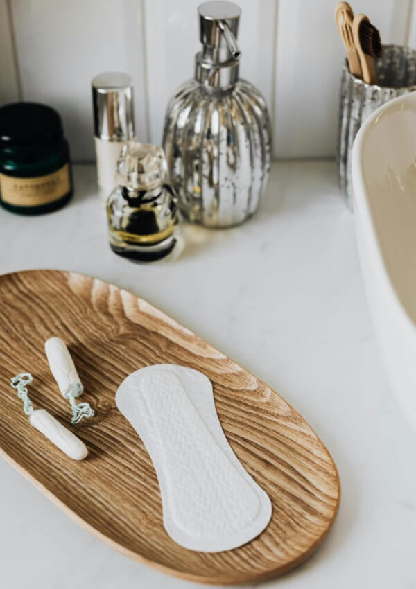 A wooden tray on a bathroom counter holds two tampons and a sanitary pad. In the background are bottles of toiletries, a jar, and a container holding toothbrushes.