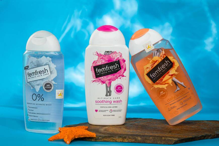 Three Femfresh intimate wash bottles are arranged against a blue background, with one bottle tilted on a wooden board and a starfish in front. Each bottle has a colorful label and distinctive design.