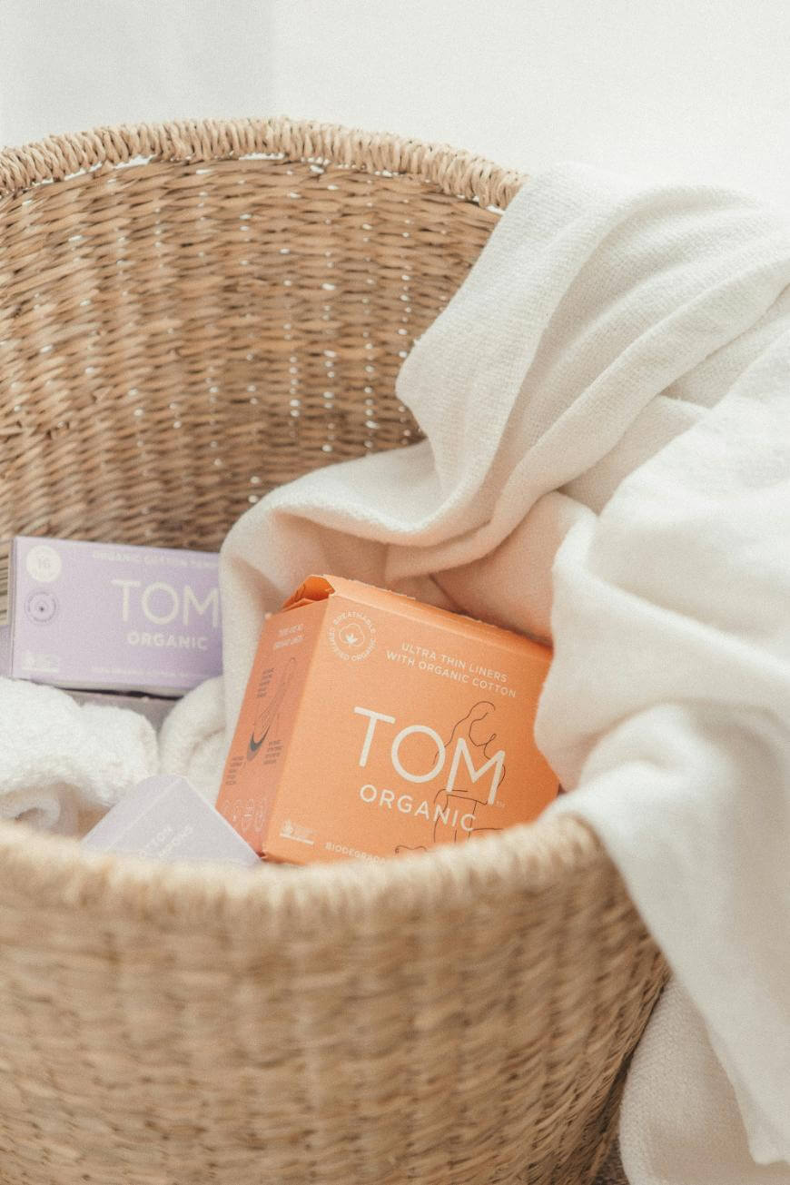 A woven basket contains white towels and boxes of TOM Organic products, including an orange box and a purple box, placed in soft, natural lighting.