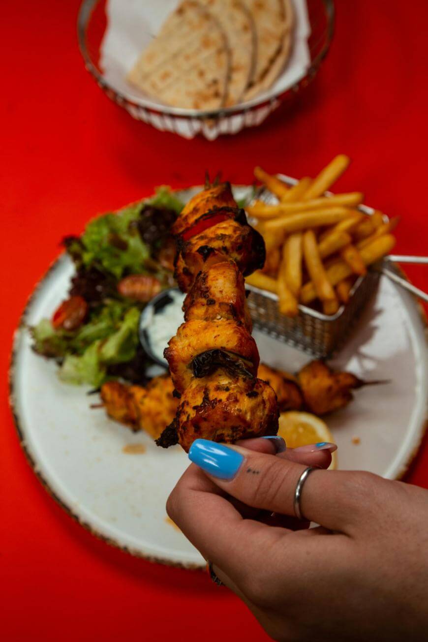 A hand with blue nail polish holds a skewer of grilled meat above a plate with fries, salad, sauce, and lemon, with a basket of flatbread in the background on a red table.