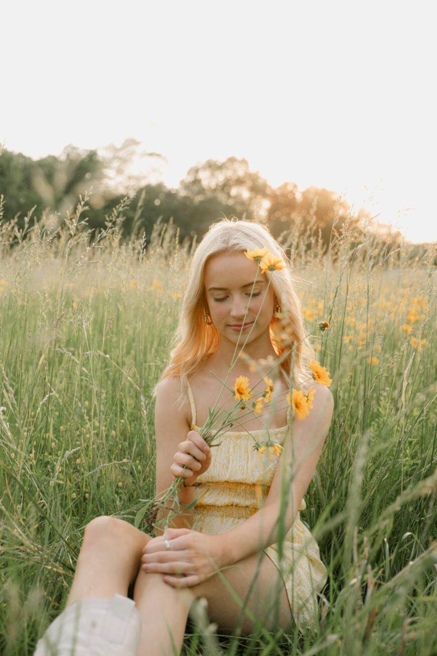 A young woman in a yellow dress sits in a grassy field at sunset, holding yellow wildflowers and smiling softly with her eyes closed. Tall grass and yellow flowers surround her.