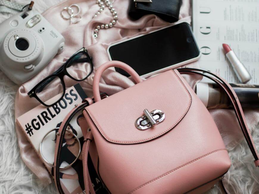 A flat lay of feminine accessories on a soft surface, including a pink handbag, glasses, a polaroid camera, lipstick, pearl necklace, rings, a phone, a wallet, and a #GIRLBOSS card.