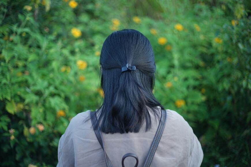 A person with shoulder-length black hair tied with a clip, seen from behind, stands outdoors in front of green bushes with yellow flowers.