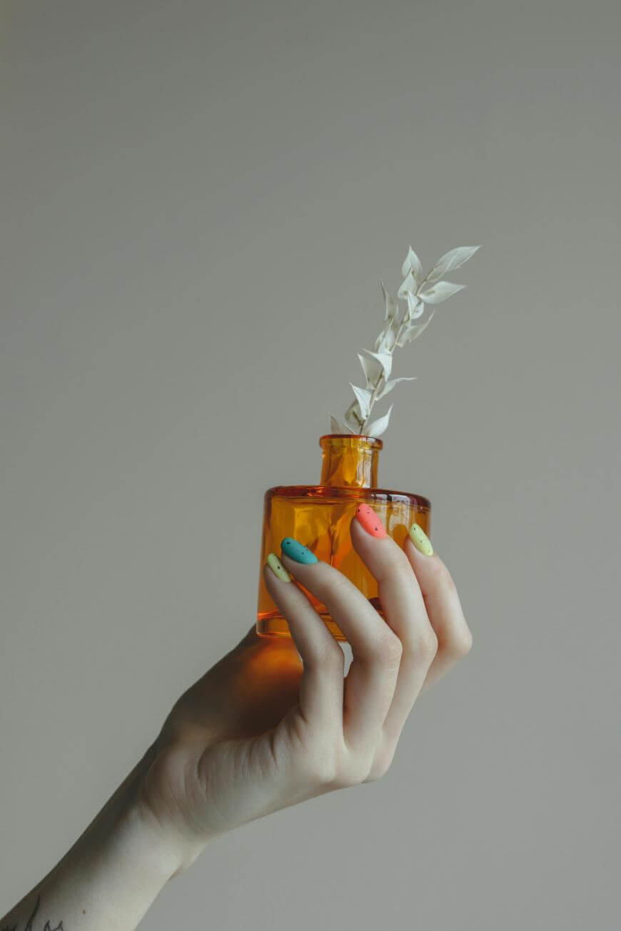 A hand with colorful, pastel-painted nails holds a small amber glass vase containing white dried leaves against a neutral background.