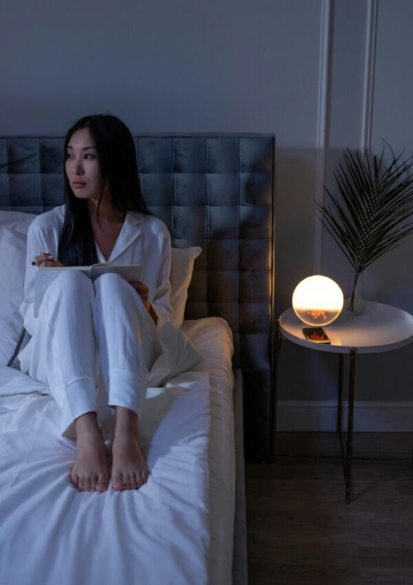A woman in white pajamas sits on a bed, holding a notebook and pen, thoughtfully reflecting as part of her night routine. A round glowing lamp and palm leaf in a vase rest on the bedside table beside her in the softly lit room.