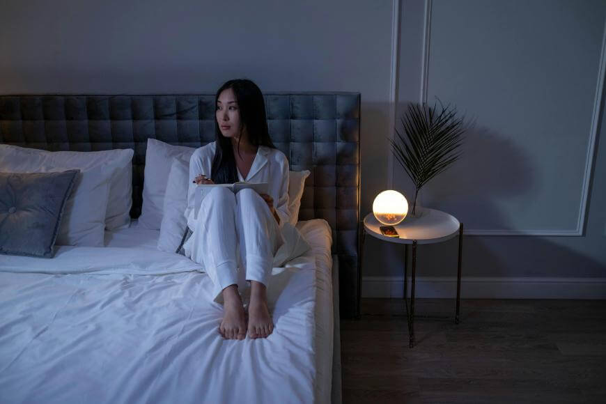 A woman in white pajamas sits on a bed at night, holding her knees and looking to the side. A round lamp and a decorative plant on a small table beside her add to the peaceful mood of her night routine.