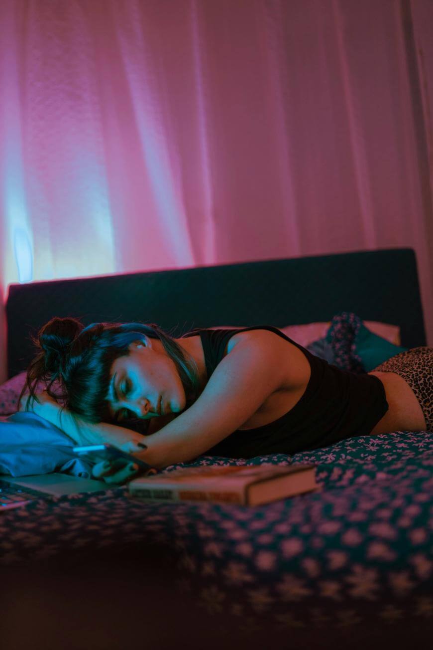 A woman lies on her side on a bed with patterned sheets, resting her head on her arm near a book and a smartphone, illuminated by soft blue and pink lighting in a dimly lit bedroom.