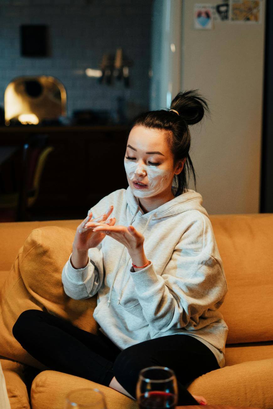A person wearing a gray hoodie sits on a couch with their legs crossed, applying white facial cream to their face in a cozy, dimly lit room.