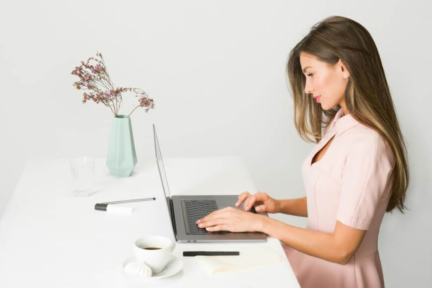 A woman in a pale pink dress sits at a white desk, typing on a laptop. There is a cup of coffee, a pen, and a mint green vase with dried flowers on the table. The background is plain white.