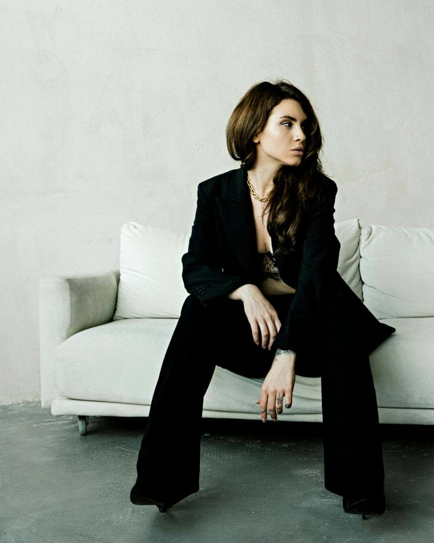 A woman with long brown hair sits on a white sofa, wearing a black suit and lace top. She looks to her left with a serious expression, one hand resting on her knee in a minimalist room with neutral tones.