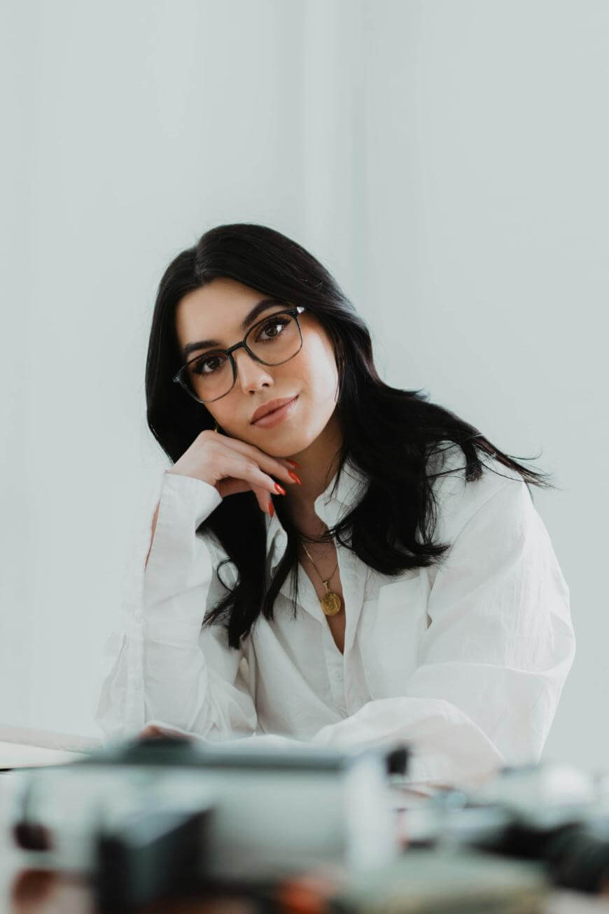 A woman with long dark hair and glasses, wearing a white shirt, rests her chin on her hand and looks at the camera. The background is bright and minimalistic.