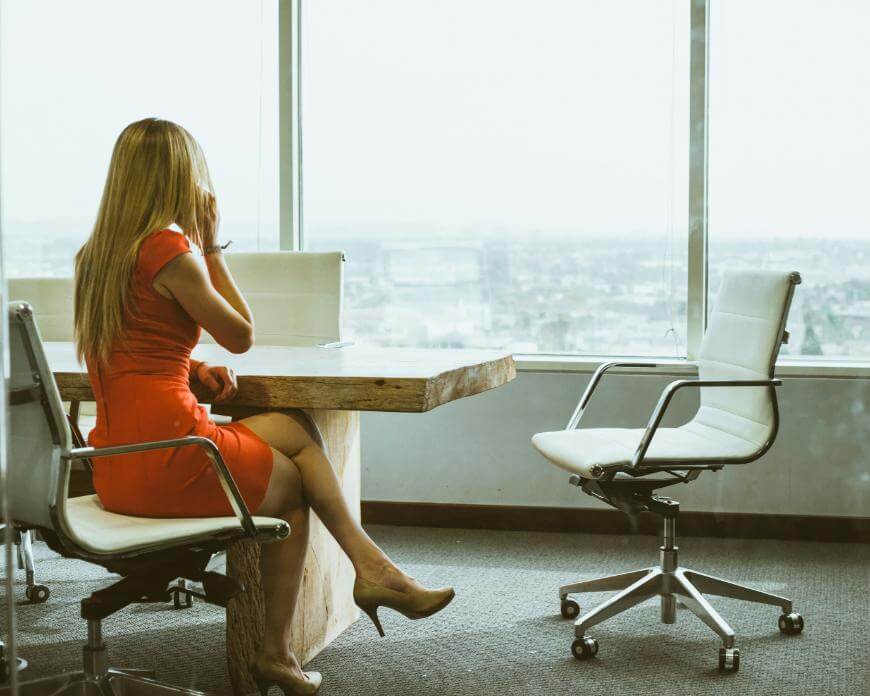 A woman in a red dress sits at a wooden conference table, looking out a large window with city views, while talking on her phone. An empty office chair is beside her.
