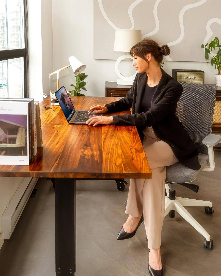 A woman in business attire sits at a wooden desk, working on a laptop in a modern, well-lit office with large windows, plants, and books.