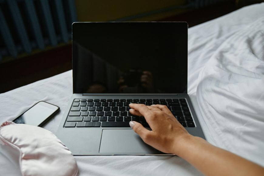 A person’s hand rests on a laptop keyboard on a bed, with a smartphone and a pink sleep mask nearby. The laptop screen is off and the bedding is white.