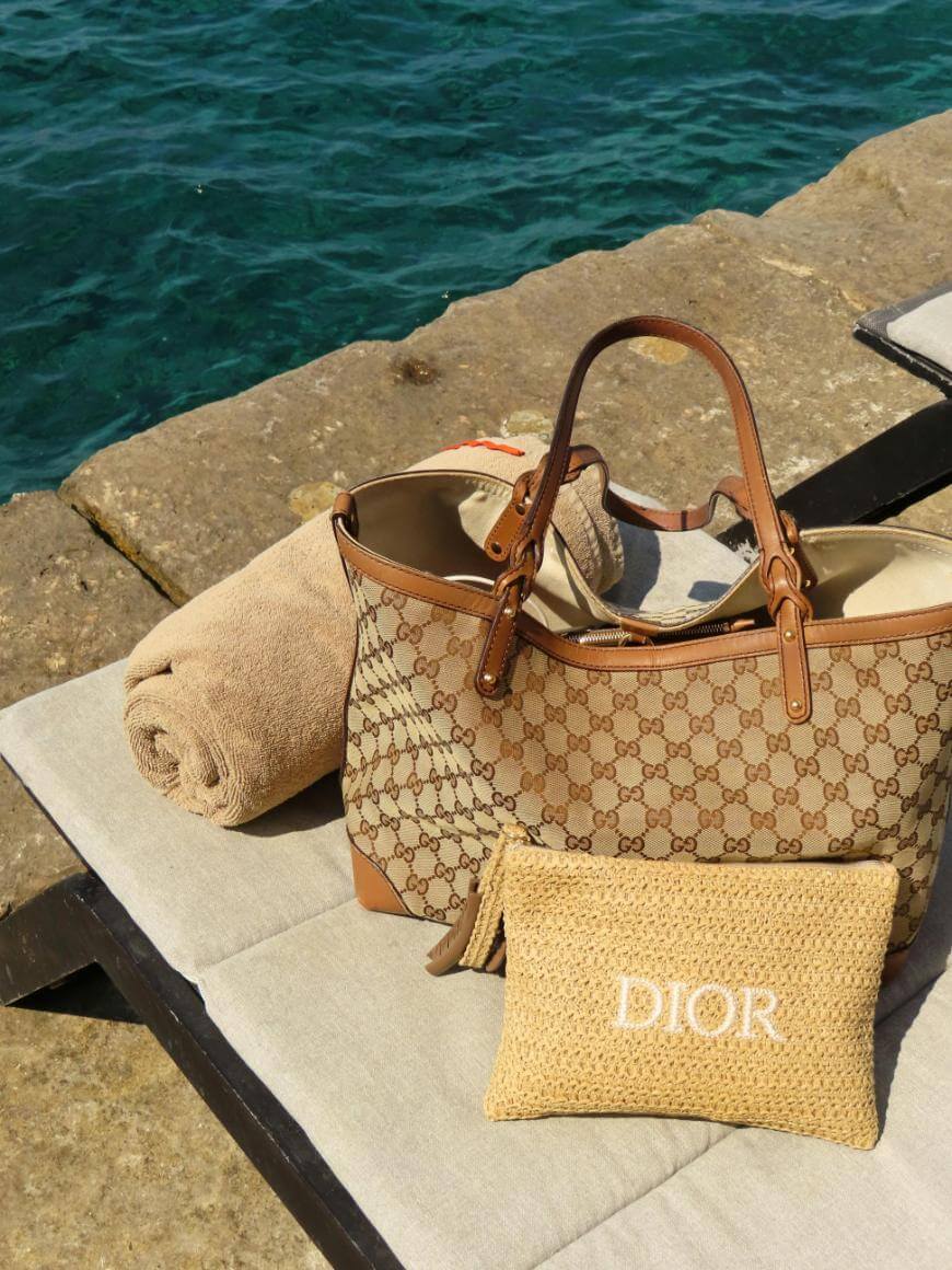 A Gucci tote bag and a woven Dior pouch rest on a cushioned lounge chair next to a rolled beige towel, with bright blue water in the background.