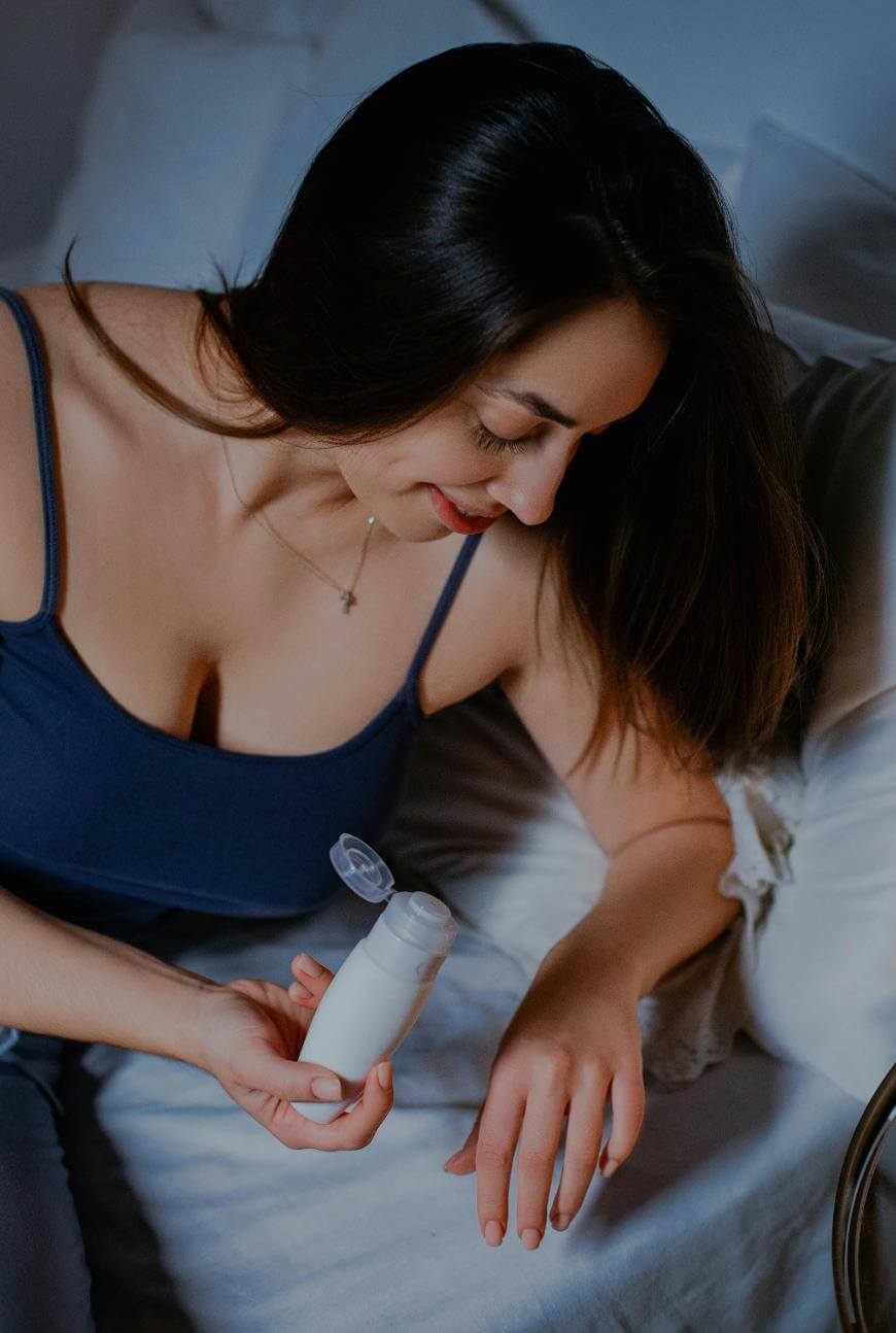 A woman in a navy tank top sits on a bed, smiling as she applies lotion from a white bottle onto her hand. She has long dark hair and is leaned slightly forward over white pillows and bedding.