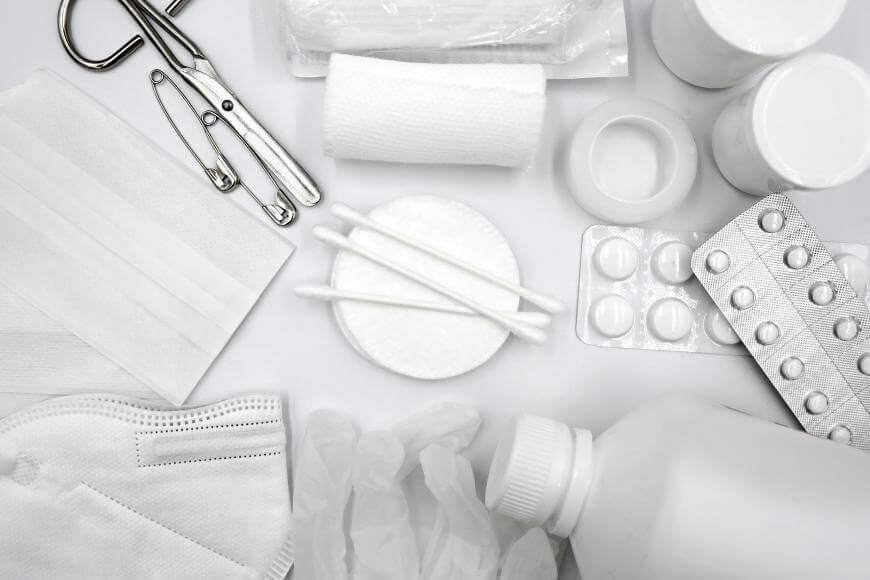 Assorted medical supplies including pills, bottles, face masks, cotton swabs, gauze, gloves, tape, and scissors are arranged on a white surface.