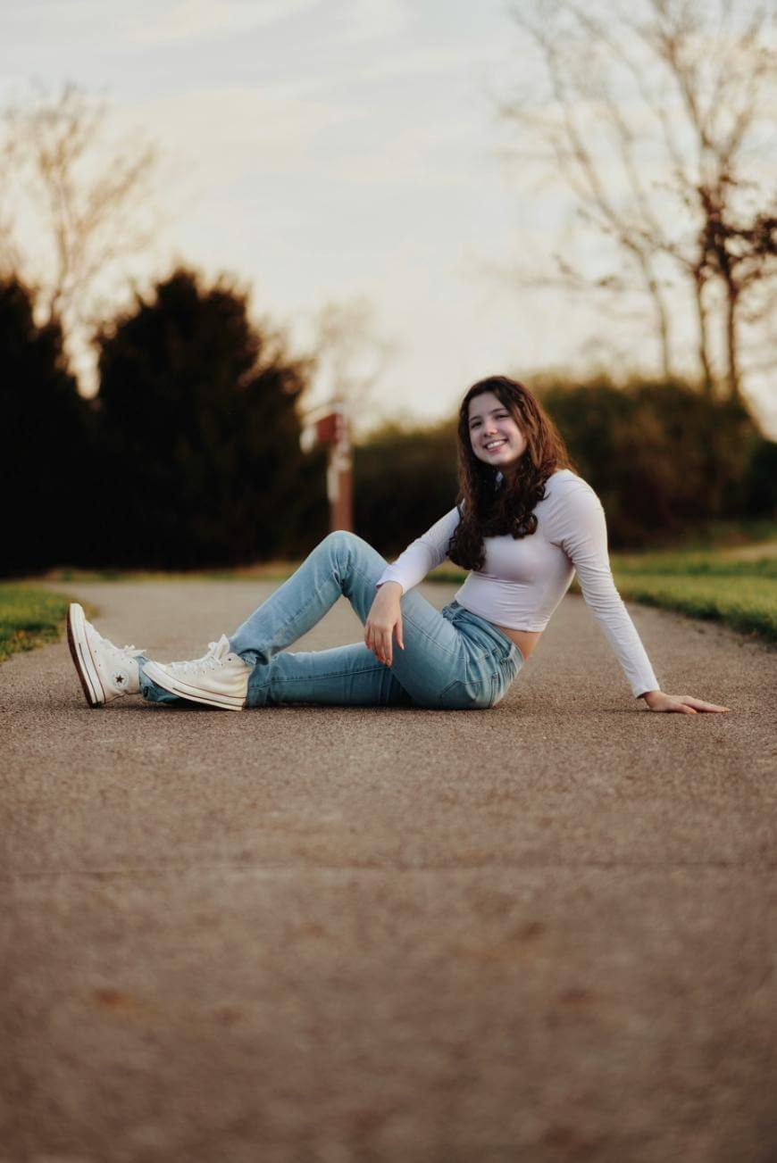 A young woman with long brown hair, wearing a white long-sleeve top, blue jeans, and white sneakers, sits on a paved path outdoors, smiling at the camera with trees and grass in the background.