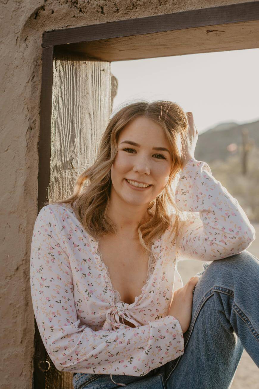 A young woman with light brown hair, wearing a white floral blouse and blue jeans, sits in a rustic wooden window frame, smiling softly at the camera with sunlight in the background.