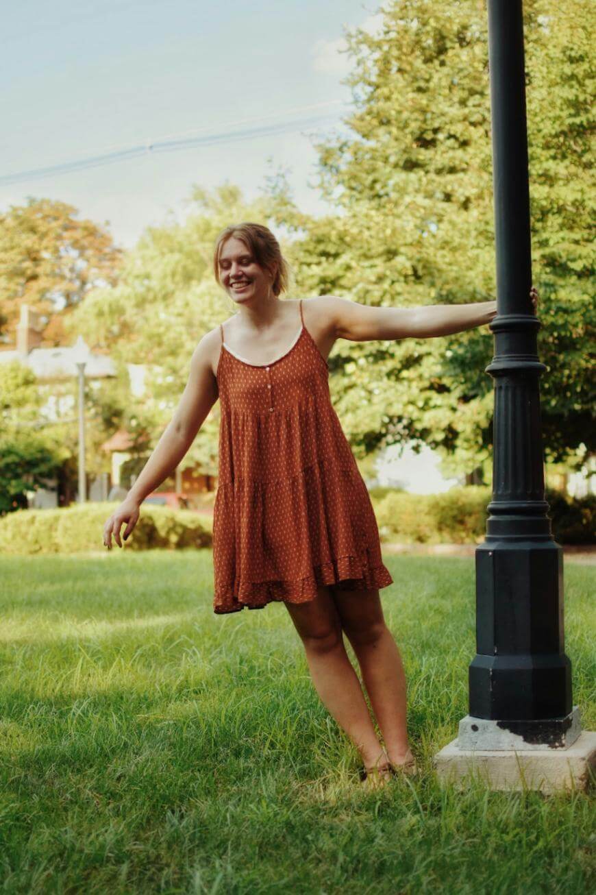 A young woman in a sleeveless rust-colored dress stands barefoot on grass, smiling and holding onto a black lamppost in a sunlit park with trees and bushes in the background.