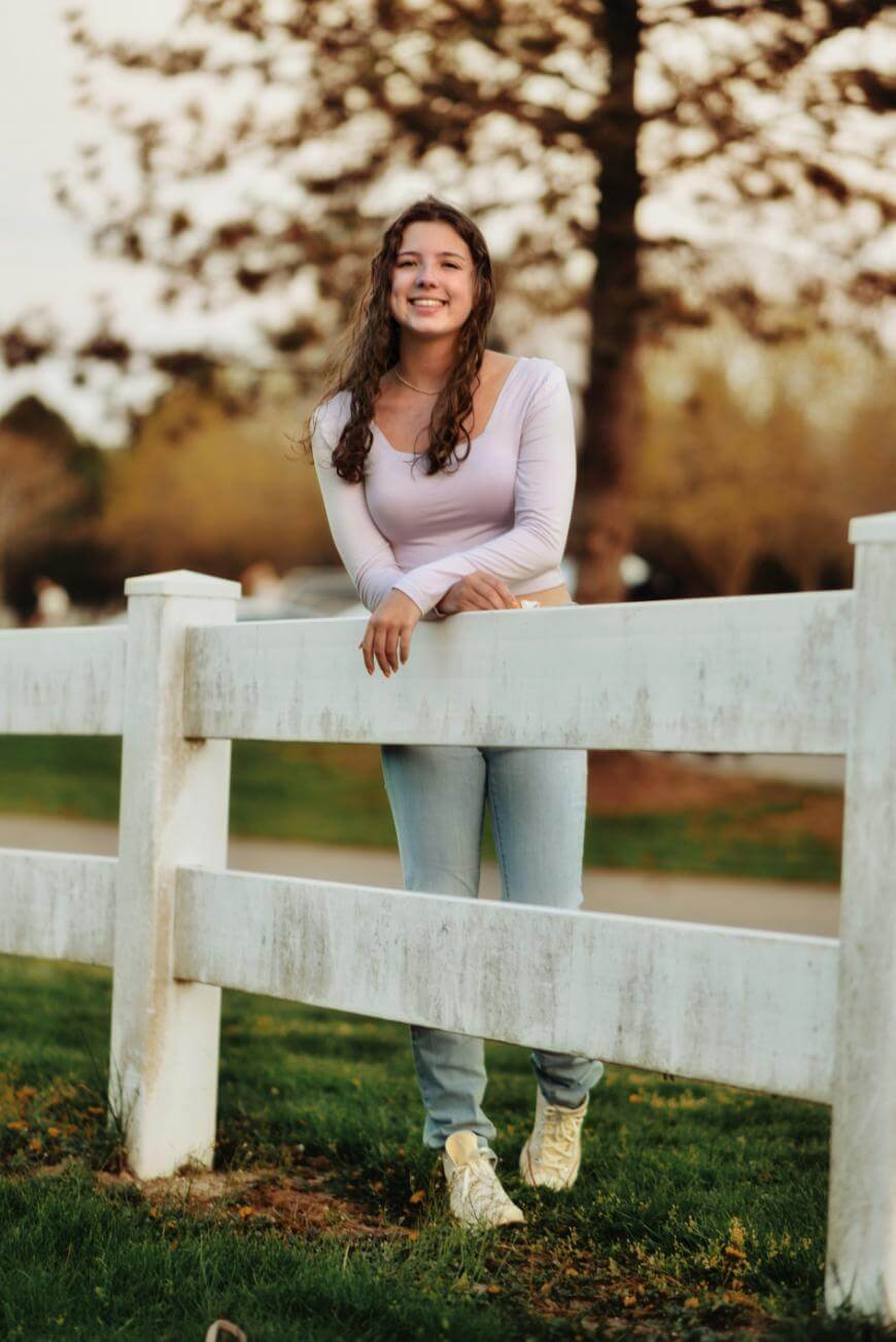 A young woman with long curly hair, wearing a light purple top and jeans, stands smiling behind a white wooden fence in a grassy outdoor setting with trees in the background.