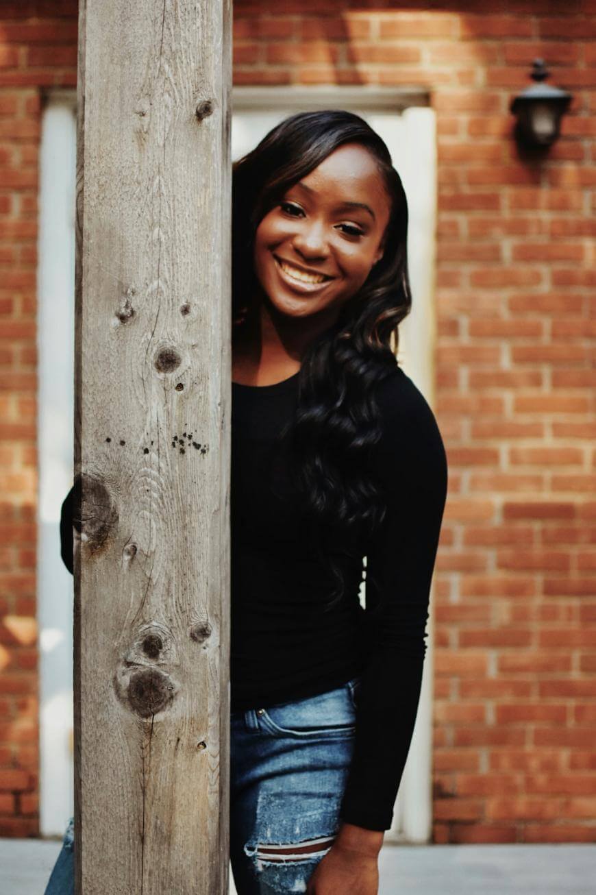 A woman with long, wavy hair wearing a black top and ripped jeans smiles while standing behind a wooden post in front of a brick wall.