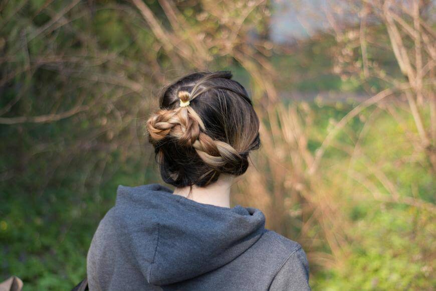 A person with brown hair styled in a braided bun, wearing a gray hoodie, is standing outdoors with their back to the camera, surrounded by greenery and bare branches.