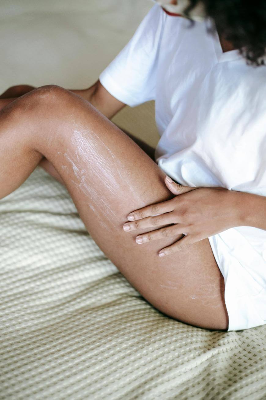 A person sitting on a bed in a white shirt applies lotion to their bare thigh, spreading it with one hand. The focus is on their legs and hand, emphasizing skin care.