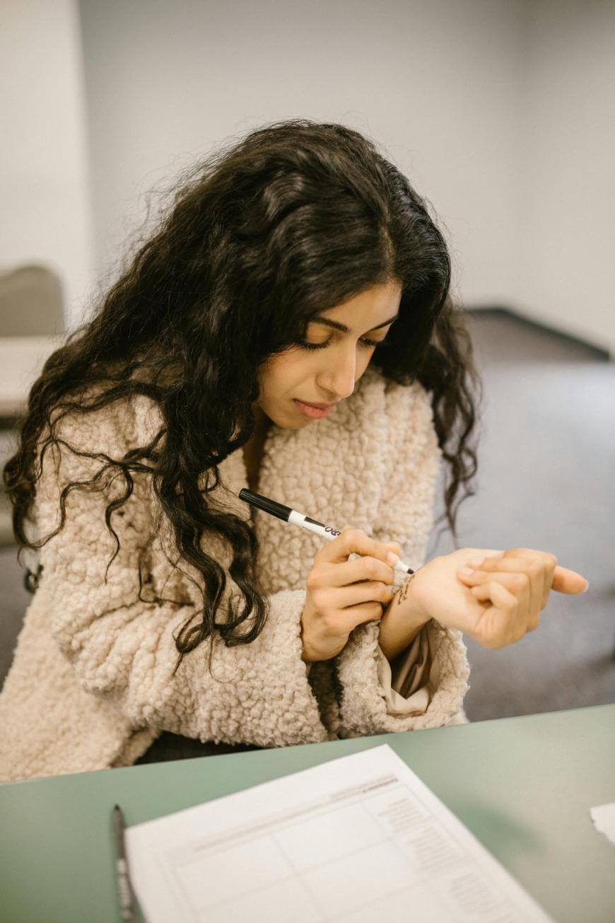 A woman with long curly hair wearing a fuzzy coat sits at a table, writing on her wrist with a pen. A document with printed text lies on the table in front of her.