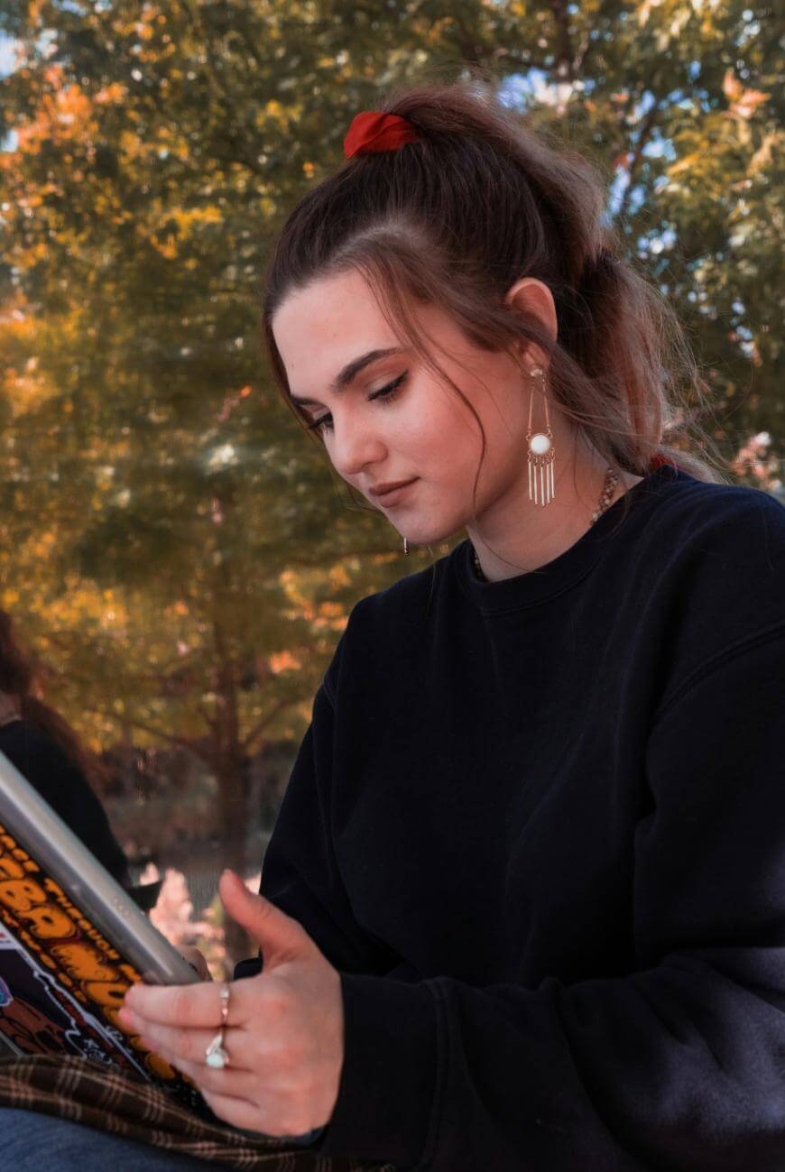 A young woman with long brown hair in a ponytail, wearing a black sweatshirt and dangling earrings, sits outdoors reading a tablet. Trees with green and orange leaves are visible in the background.