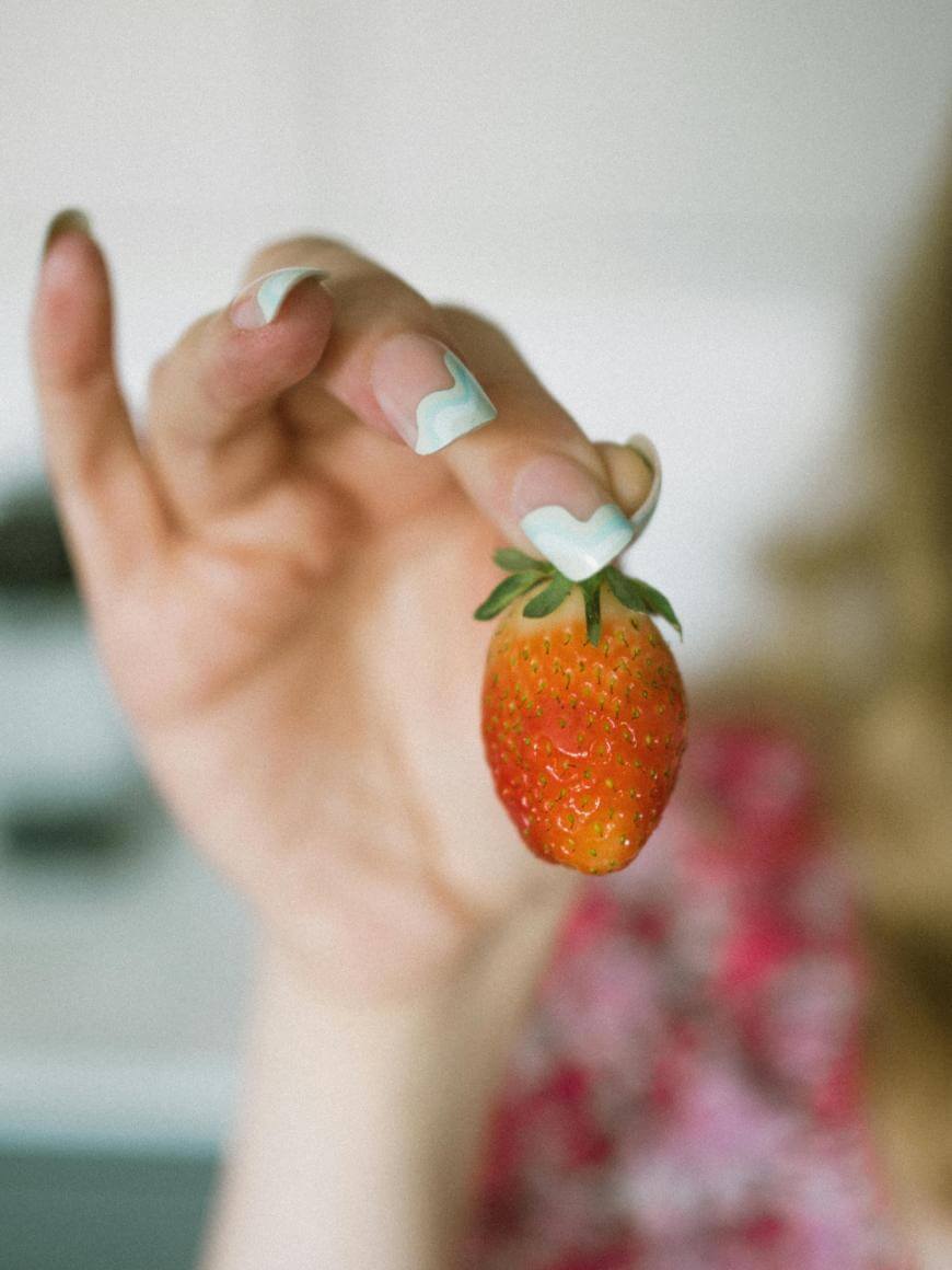 A hand with long, decorated nails holds a ripe strawberry by its green stem, with the background softly blurred.