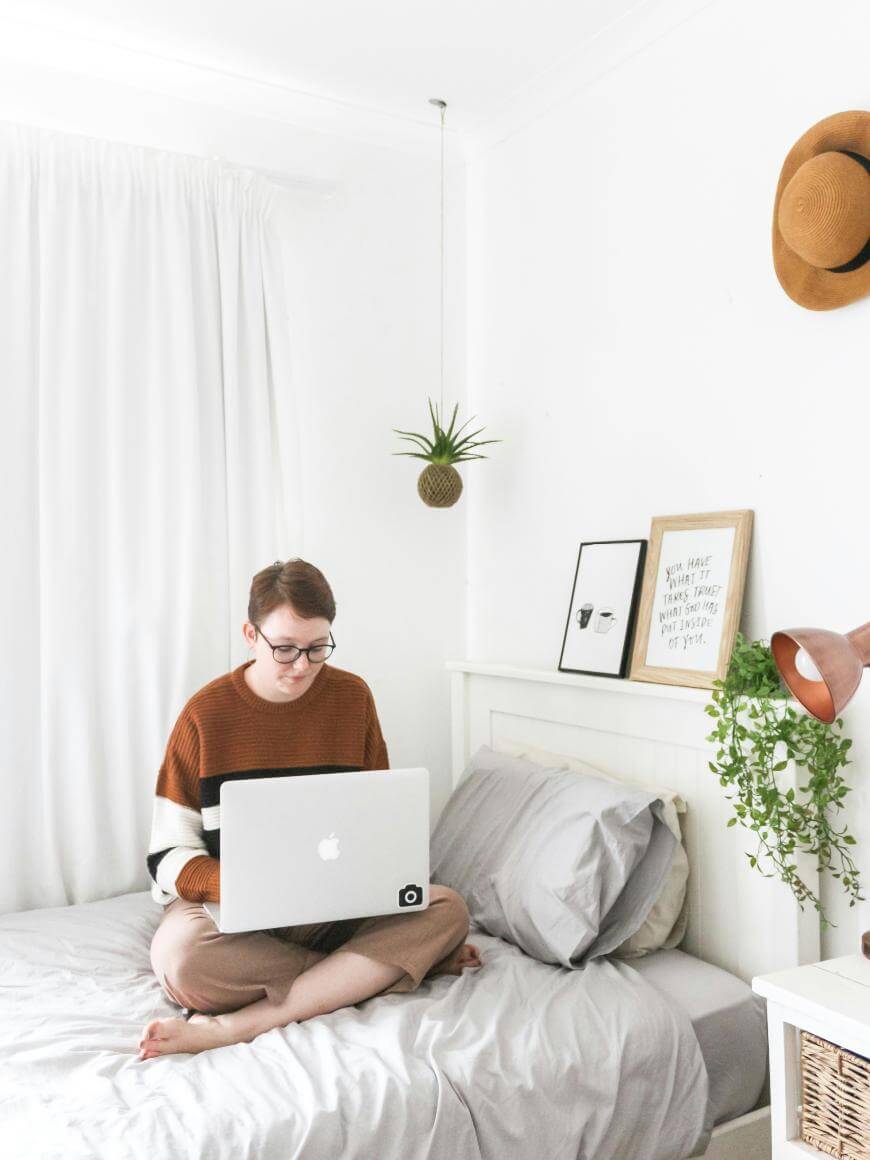 A person wearing glasses and a brown sweater sits cross-legged on a neatly made bed using a laptop in a bright, white bedroom decorated with plants, framed art, and a brown hat on the wall.