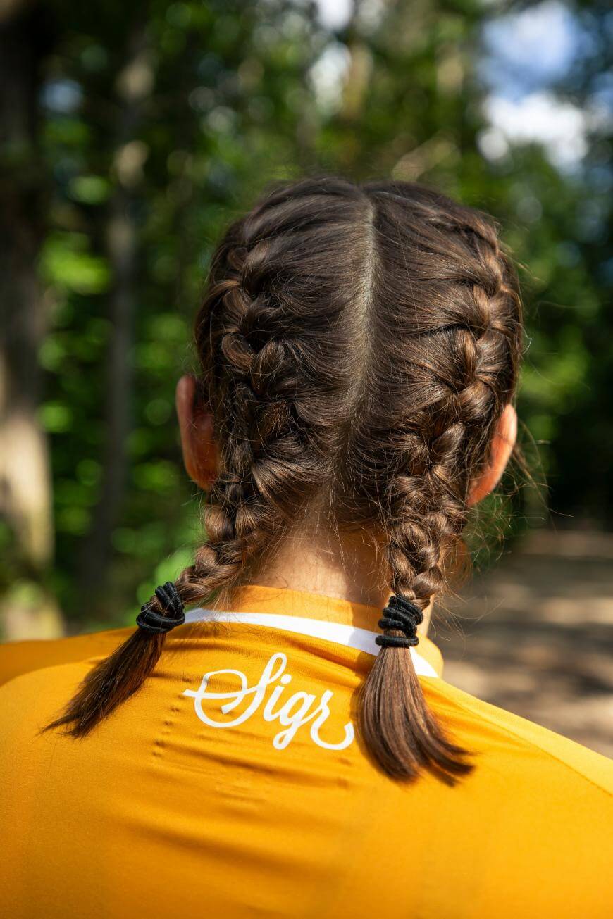A person with two neatly braided pigtails, seen from behind, wears a yellow shirt with “Sigr” written in white cursive letters on the collar. The background shows a sunlit, green forest.