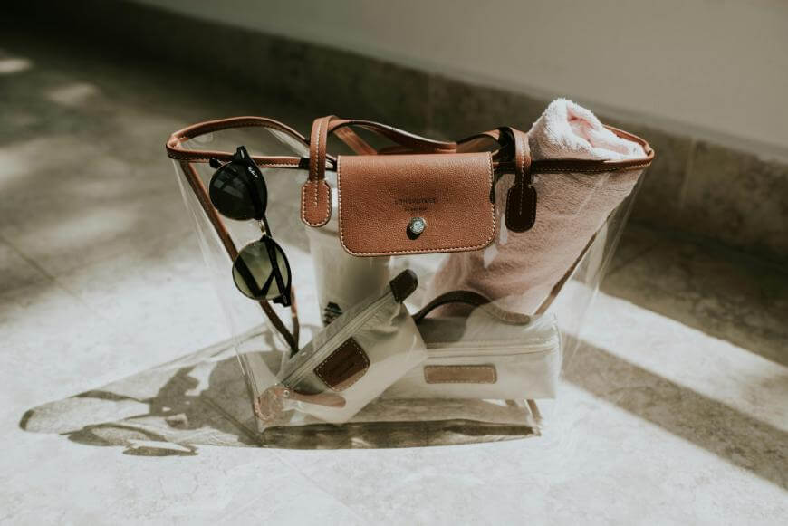 A clear tote bag with brown leather trim sits on a sunlit floor. Inside are a rolled pink towel, a small pouch, and a zippered pouch. A pair of black sunglasses hangs from one of the bag’s handles.