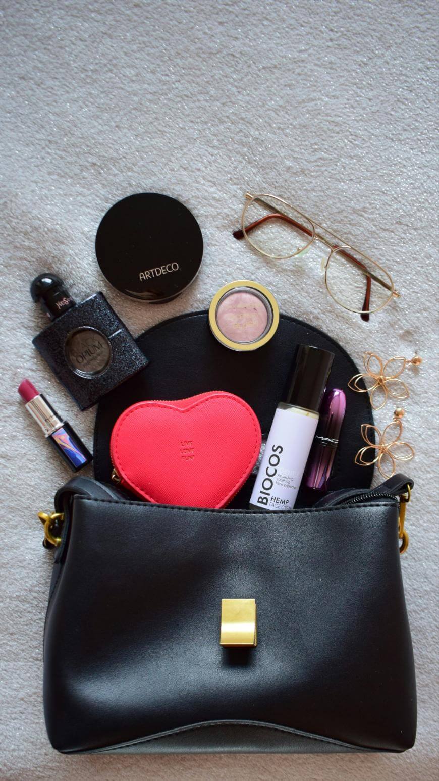 A black purse with a gold clasp lies open, displaying contents: glasses, earrings, lipstick, perfume, powder, blush, face mist, heart-shaped case, and a small black pouch on a light fabric background.