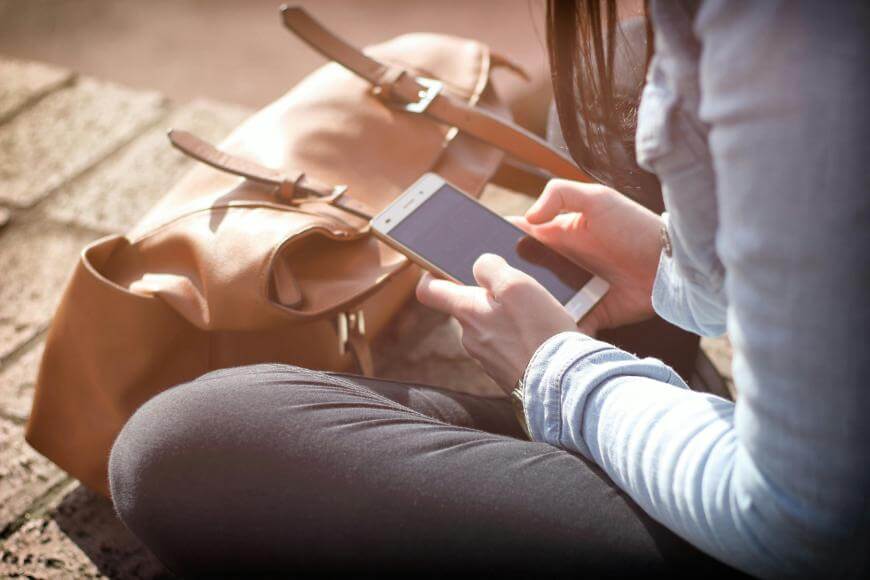 A person sitting outdoors, holding and using a smartphone. A brown handbag rests on the ground beside them. The person is wearing dark pants and a light blue long-sleeve shirt.