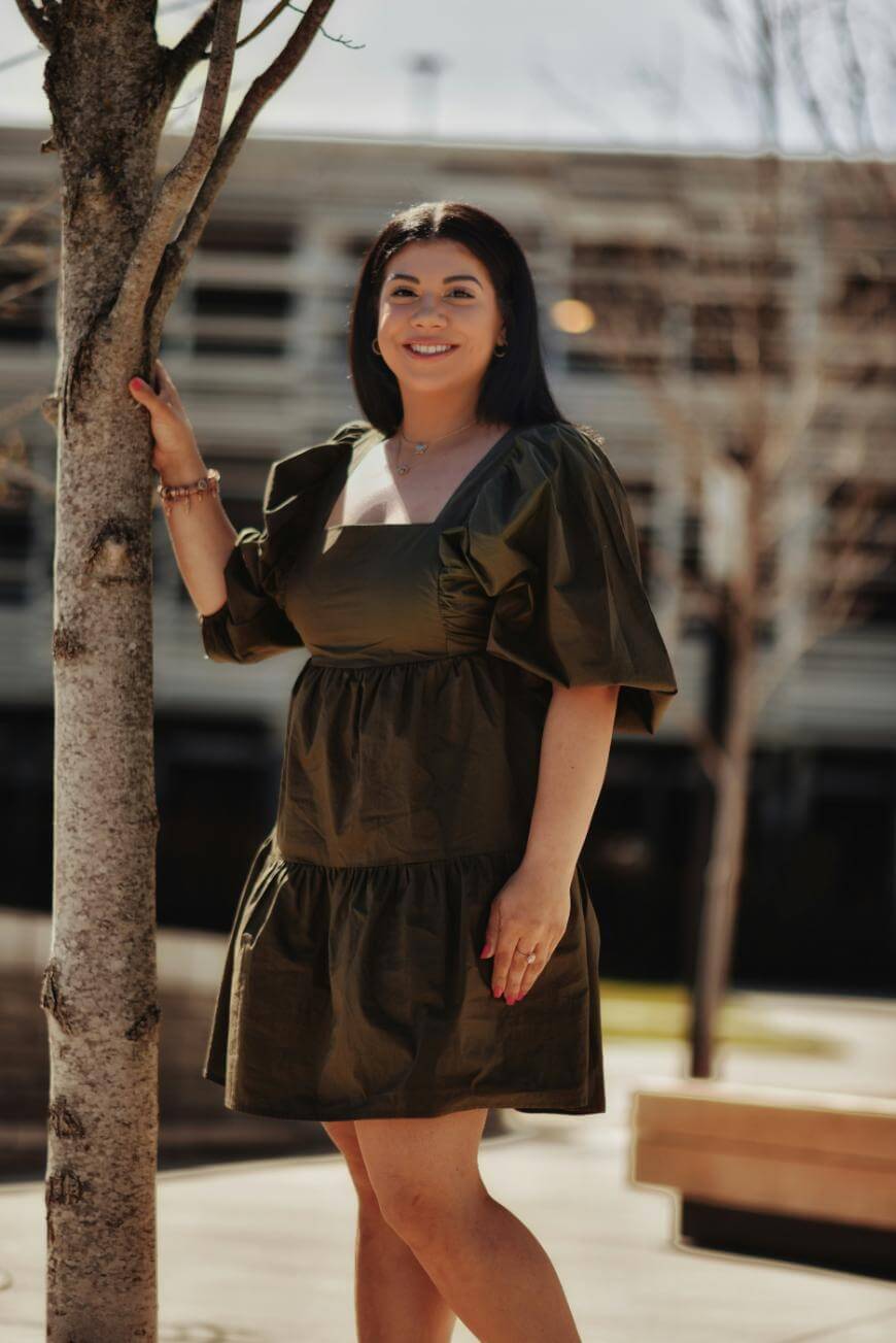 A woman in a dark green, puff-sleeve dress stands outdoors next to a tree, smiling at the camera. The background features blurred buildings and bare trees, suggesting a sunny day in early spring or autumn.