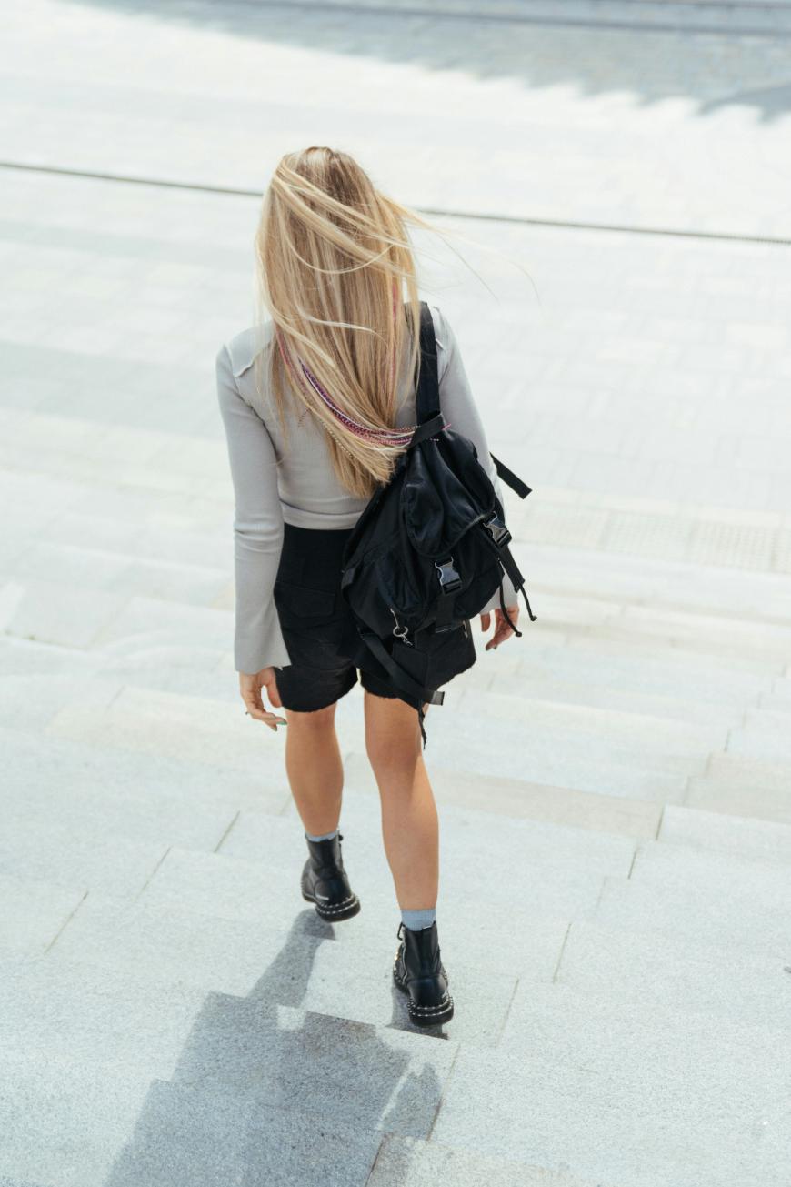 A young woman with long blonde hair, wearing a light grey top, black shorts, black boots, and a black backpack, walks down outdoor stone steps on a sunny day.