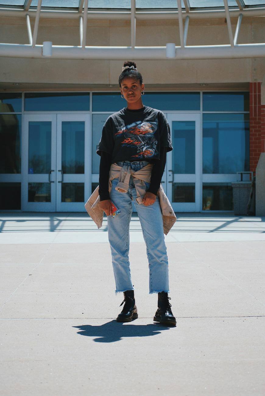 A person stands confidently outside a building with glass doors, wearing a black graphic T-shirt, ripped jeans, a jacket tied around their waist, and black boots.